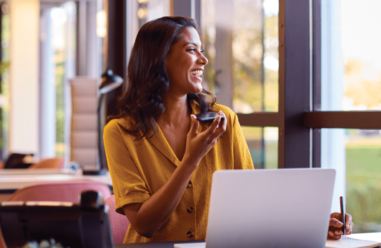 Mature Businesswoman Working On Laptop At Desk In Office Talking Into Mic Of Mobile Phone