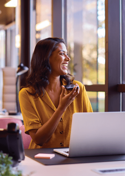 Mature Businesswoman Working On Laptop At Desk In Office Talking Into Mic Of Mobile Phone