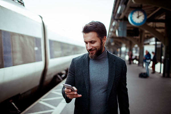 Mann schaut und lächelt auf sein Handy, während er in einem Bahnhof am Zug entlanggeht.