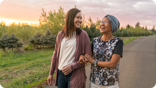 A senior woman with cancer walks with her adult daughter at sunset down a rural road.