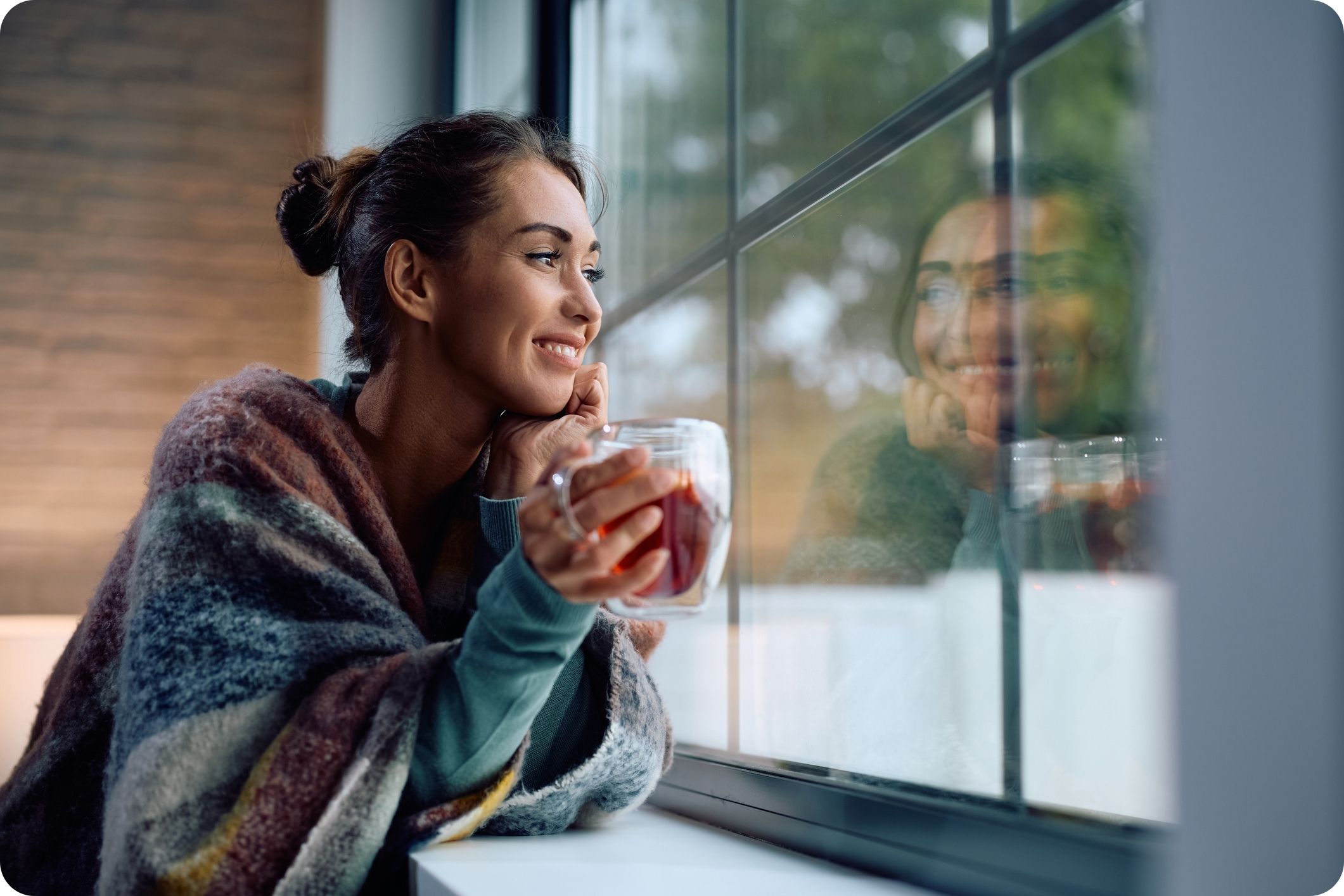 Smiling woman enjoying a cup of tea while looking out of a window at home.