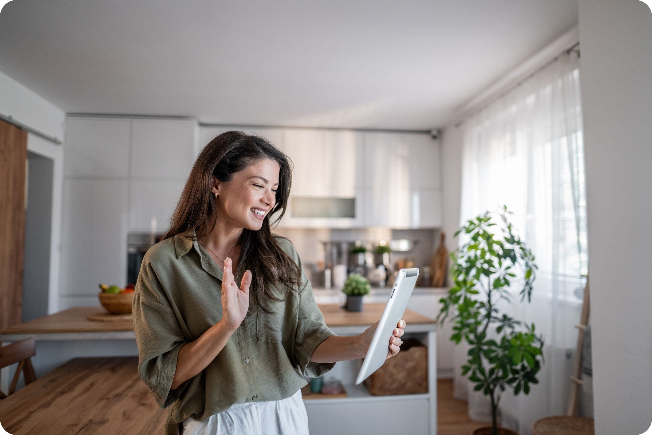 Smiling woman in a modern kitchen waving at a tablet during a video call with family or friends