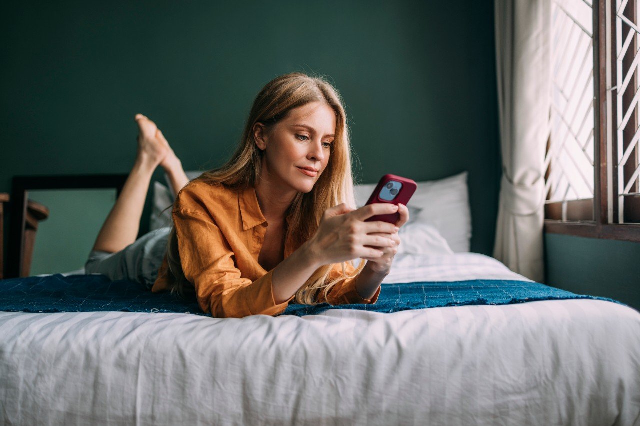 Smiling woman lying on a cozy bed, chatting on her mobile phone.