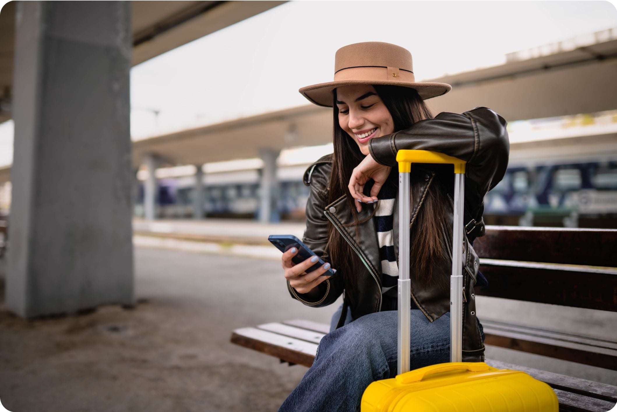 Young woman smiling while sitting on a subway platform with a suitcase, ready for travel or commuting.