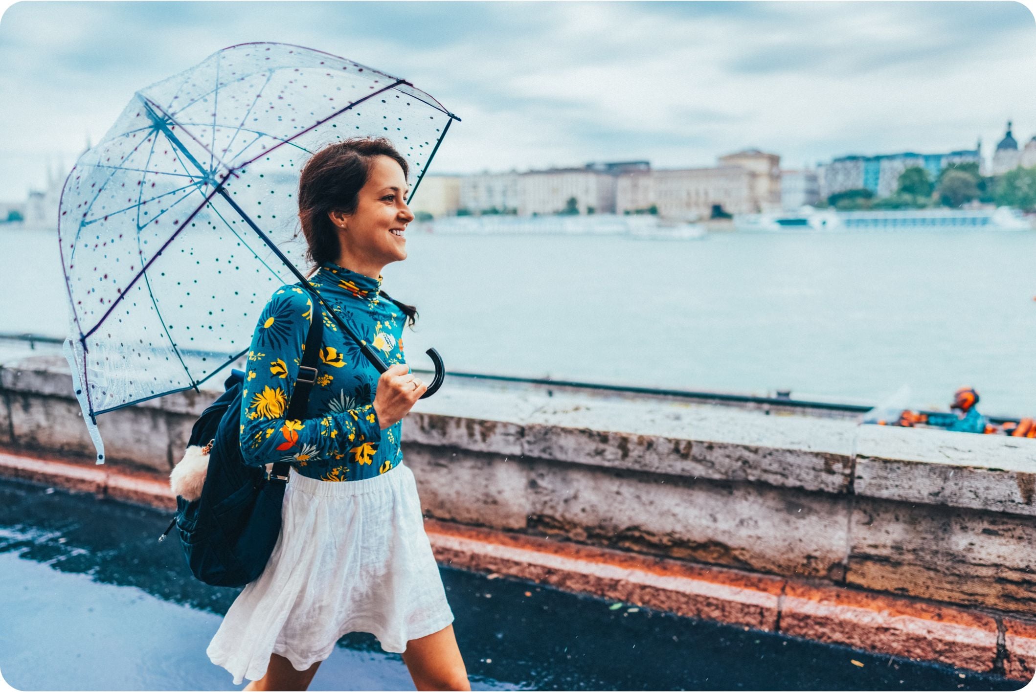 A female expat walks along a riverside path on a rainy day, holding an umbrella and looking ahead as she passes the water's edge.