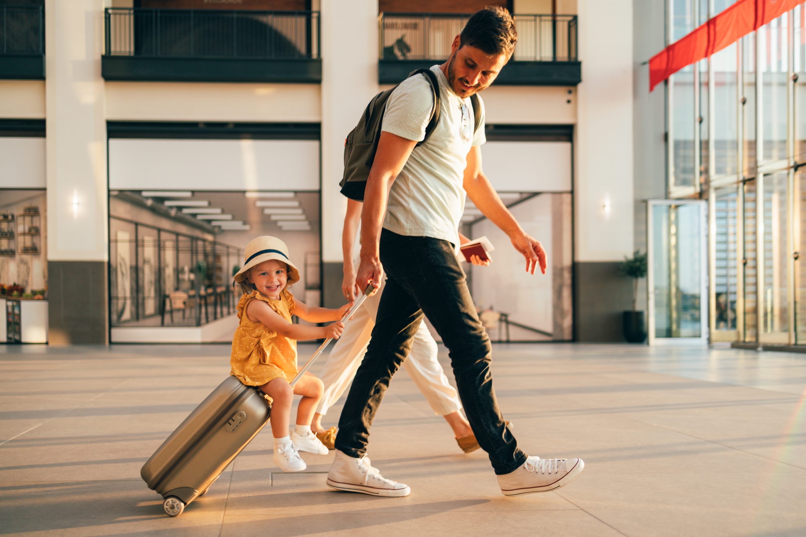 Cheerful husband and his wife walking through the airport with their young daughter sitting on a suitcase.