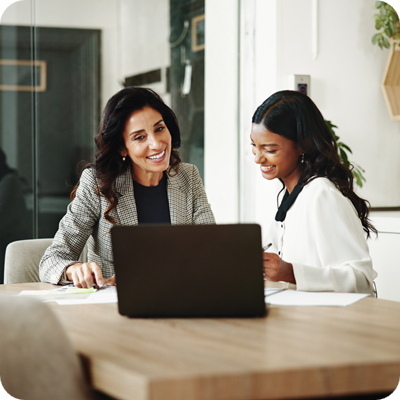 Two women sit at a table with a laptop in front of them.