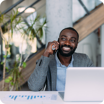 Portrait of handsome afro-american businessman sitting at desk in his office working on laptop and talking on a mobile phone.