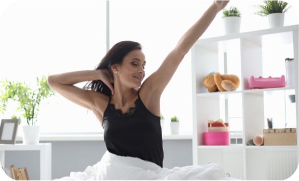 A woman stretching in bed in a bright room beside shelves