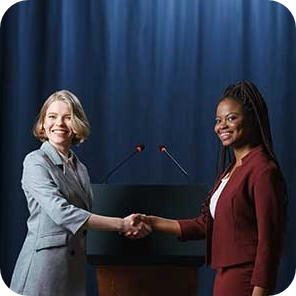 Two young female diplomats in professional attire shaking hands with warm smiles, symbolising cooperation and diplomacy.