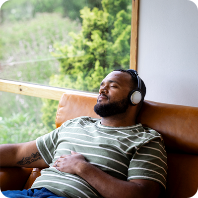 A mid adult man listening to music through wireless headphones and sitting back on a leather sofa in his living room at home