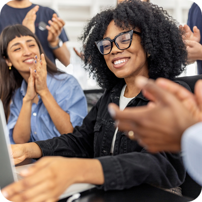African woman sitting at desk shares good news with her team clapping and celebrating a business achievement in a lively office