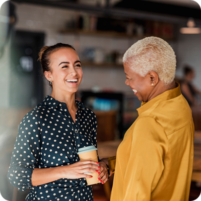 Two women colleagues laughing while standing in a cafe at their workplace.