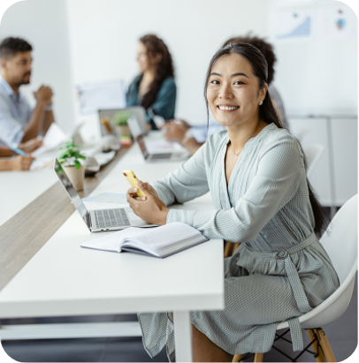An Asian businesswoman sitting in a meeting room, looking at the camera, with colleagues working in the background.