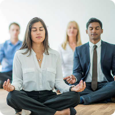 A multi-ethnic group of young business men and women in semi-casual office clothes are sitting on the floor and meditating to relax in an indoor, sunlit office.
